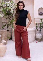 Woman wearing a black sleeveless high neck top and rust pants standing in an outdoor setting with plants and decorative pots.