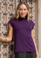 Woman wearing a sleeveless purple high neck top standing against a wooden door.