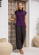 Woman wearing a purple sleeveless top and black pants standing in front of a wooden door.