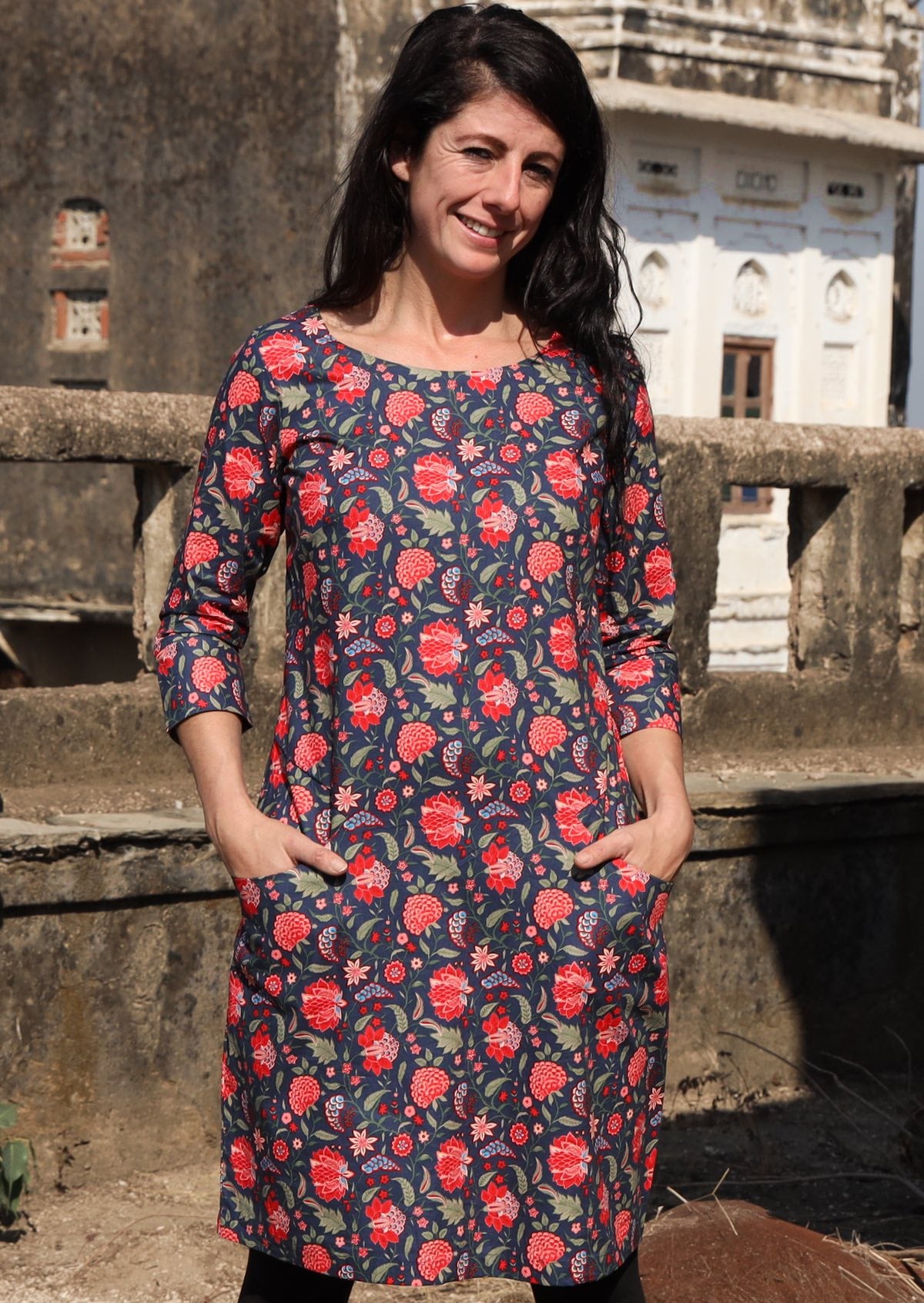 Woman with hands in pockets wearing 100 percent long sleeve cotton dress with deep blue base and raspberry red floral print 