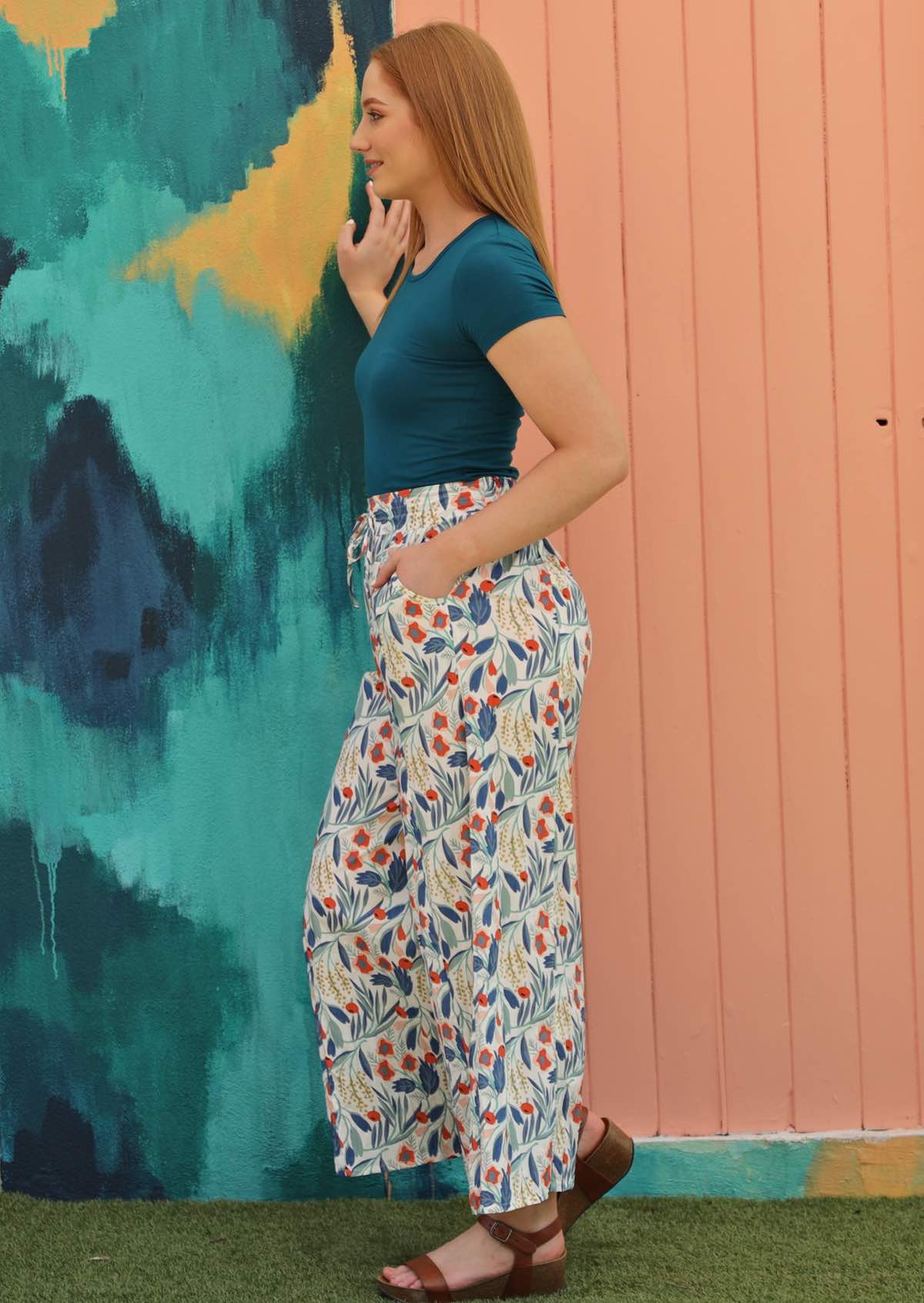 Model wears floral pants with a white base styled with a short sleeve top and sandals. 