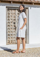 Woman in a white cotton dress with hands in pockets standing in front of a wooden door.