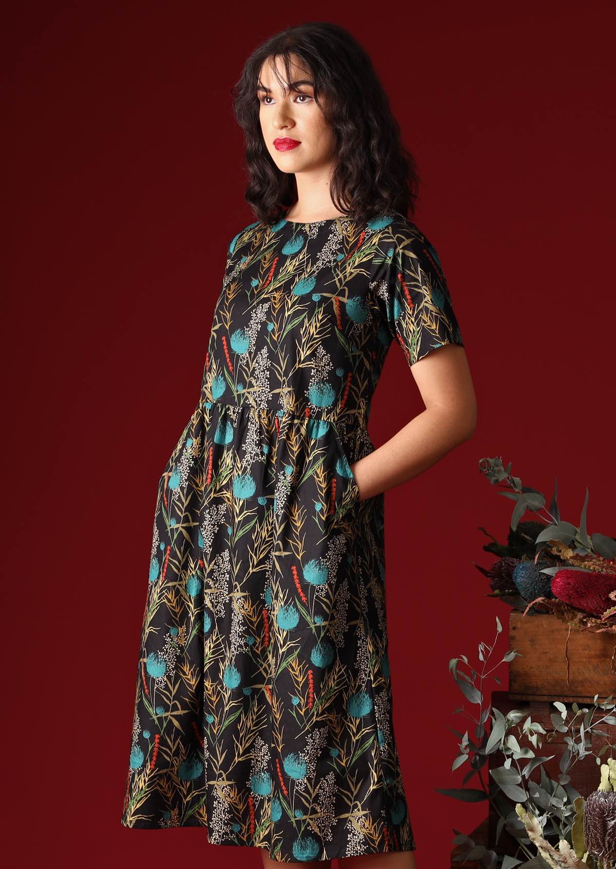 Model wears a 100% cotton floral black base dress with pops of blue and red. This loose fitting, below knee length dress has short sleeves, a high round neckline and pockets.