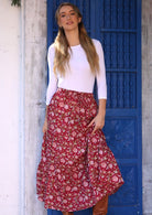 Model standing by blue door wears 100% cotton maxi skirt with pink floral print on red cranberry base.