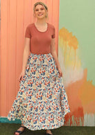 Model wears a flowing maxi skirt with green, blue, red and yellow florals on a white base. 