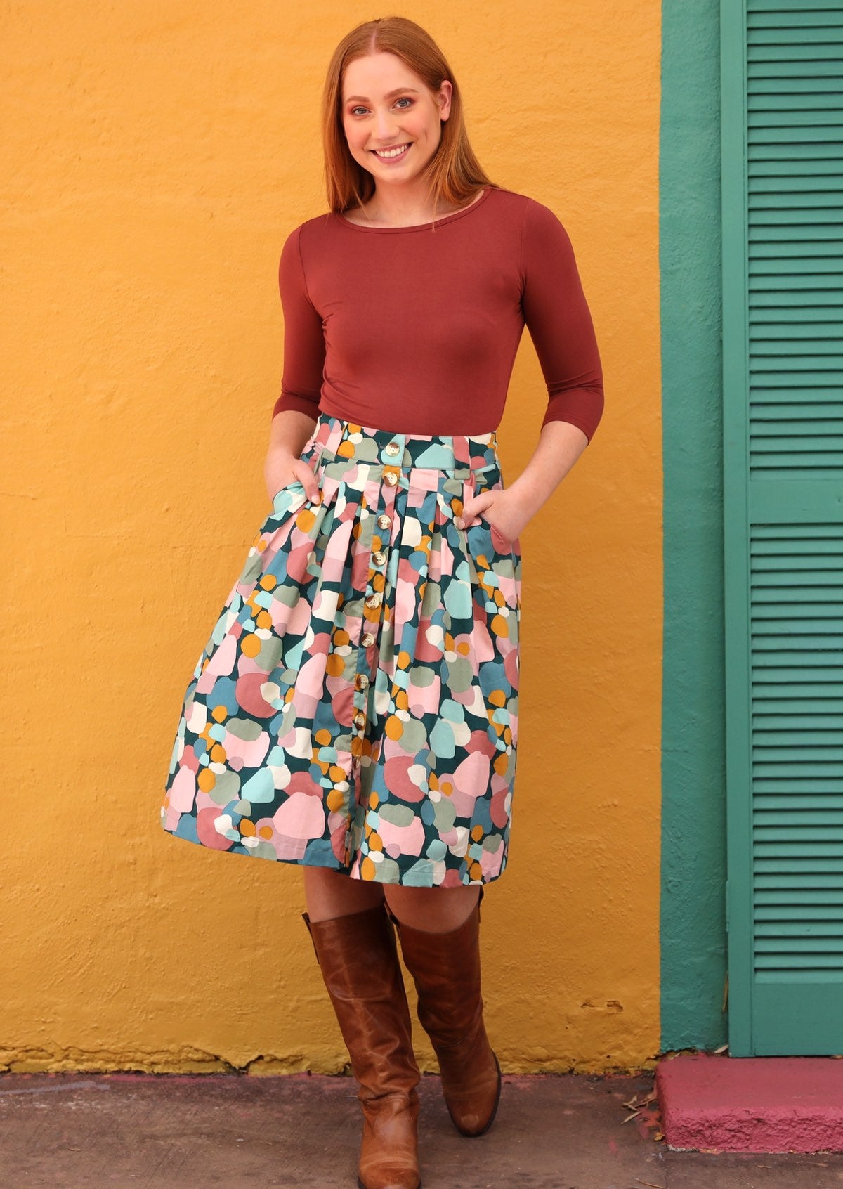 Woman wearing a pastel print knee length A-line skirt paired with tan boots and a rust top, with hands in pockets