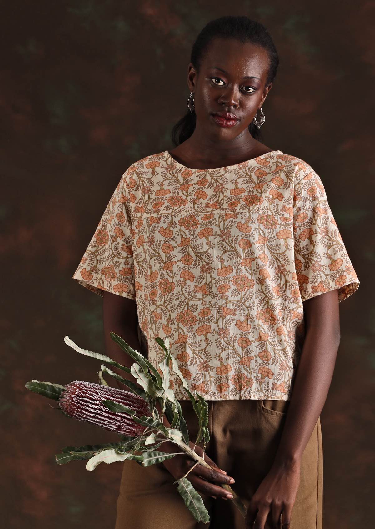 Model wears boxy cotton short sleeve top with a peachy pink floral print on a cream base. Top has a round neckline and decorative buttons down the centre of the back.