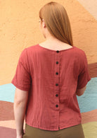Model wears a peachy terracotta top with non functional, decorative buttons on the back. 