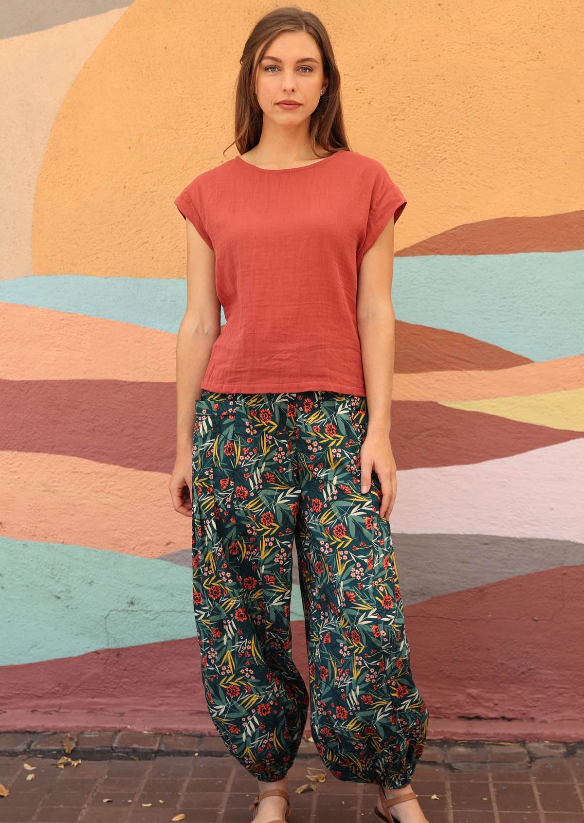 Woman wearing green cotton floral harem pants with pink top standing in front of sun mural