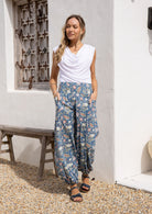 Woman wearing a white rayon top and a dusty blue floral pants with hands in deep pockets.