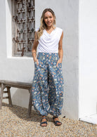 Woman wearing a white top and blue floral pants with wide elastic waist and ankles standing in front of a white wall with a wooden bench.
