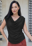 Woman wearing a black sleeveless top with a draped neckline, standing against a patterned wall.