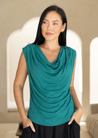 Woman wearing a jade green sleeveless top with a draped neckline against a neutral background, with hands in pockets of pants