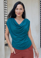 Woman wearing a teal blue sleeveless top with a draped cowl neckline with pleats in shoulders, and rust pants