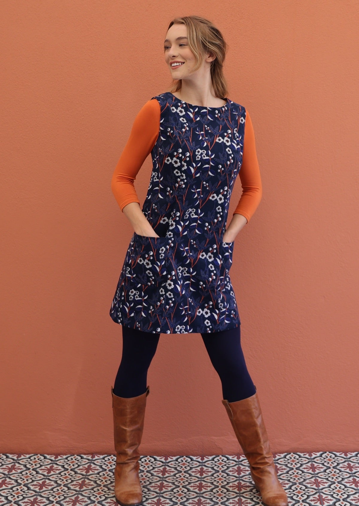 Woman poses in cotton corduroy sleeveless tunic layered over navy leggings and orange long sleeve top, with hands in pockets
