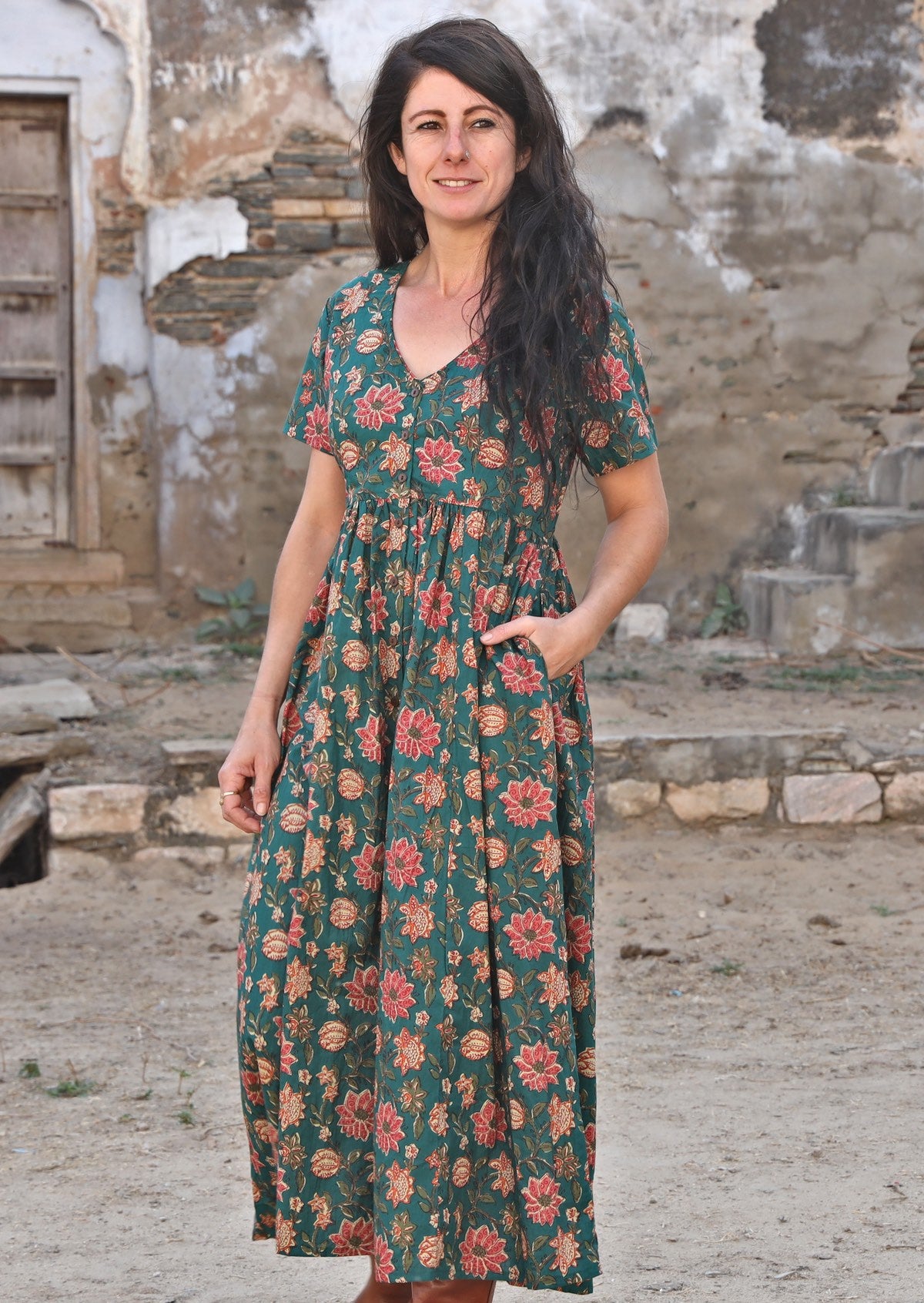 Model standing slightly sideways and smiling with one hand in pocket wearing 100 percent cotton dark jade green button through v-neck dress with pockets