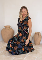 Model sitting in a retro inspired knee length cotton dress with piped detailing in black with orange and pink florals.