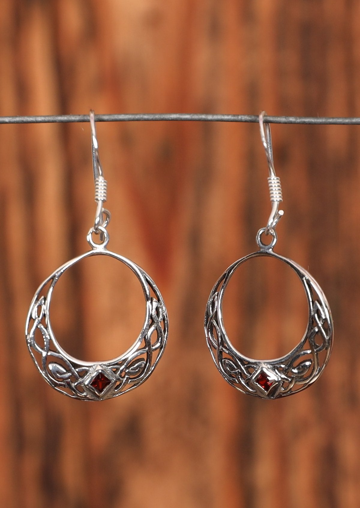 92.5% silver hook earrings in the shape of a woven moon with a red diamond shaped gem set at the base. Hanging on a wire for display.