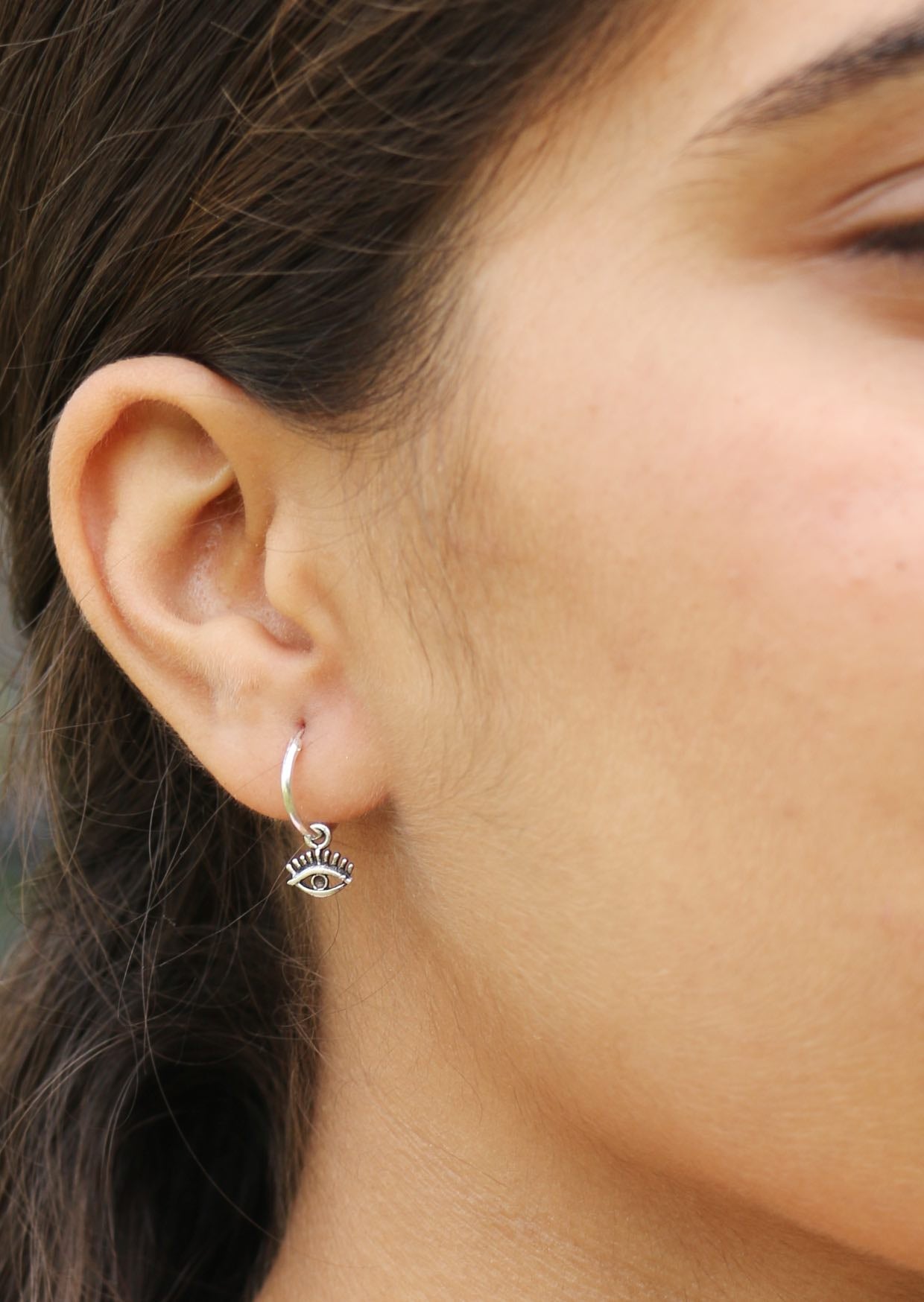 Close up image of a person wearing a silver hoop Mystical Eye earrings with dangling eye-shaped charms. 