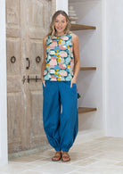 Woman wearing a colourful sleeveless top and blue pants standing in front of wooden doors.