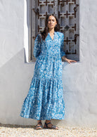Woman in a blue floral maxi dress, 100 percent cotton with sleeves, standing in front of a white wall with a decorative metal grille.