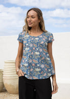 Woman wears dusty blue based floral cotton cap sleeve top with pin tuck detail across the bust and a scoop neckline