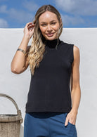Woman wearing a black sleeveless top and blue pants against a white wall with a blue sky.