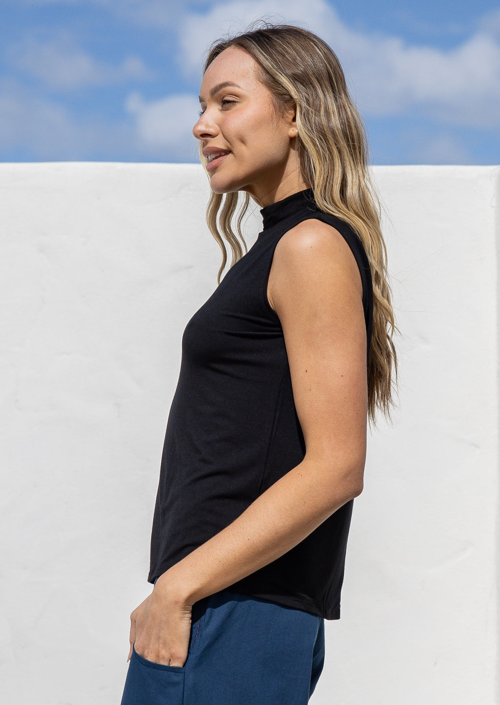 Woman wearing a black sleeveless top with a high neckline against a white wall with blue sky.