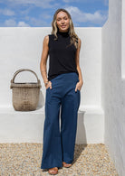 Woman in black sleeveless top and blue pants standing in front of a white wall with blue sky.