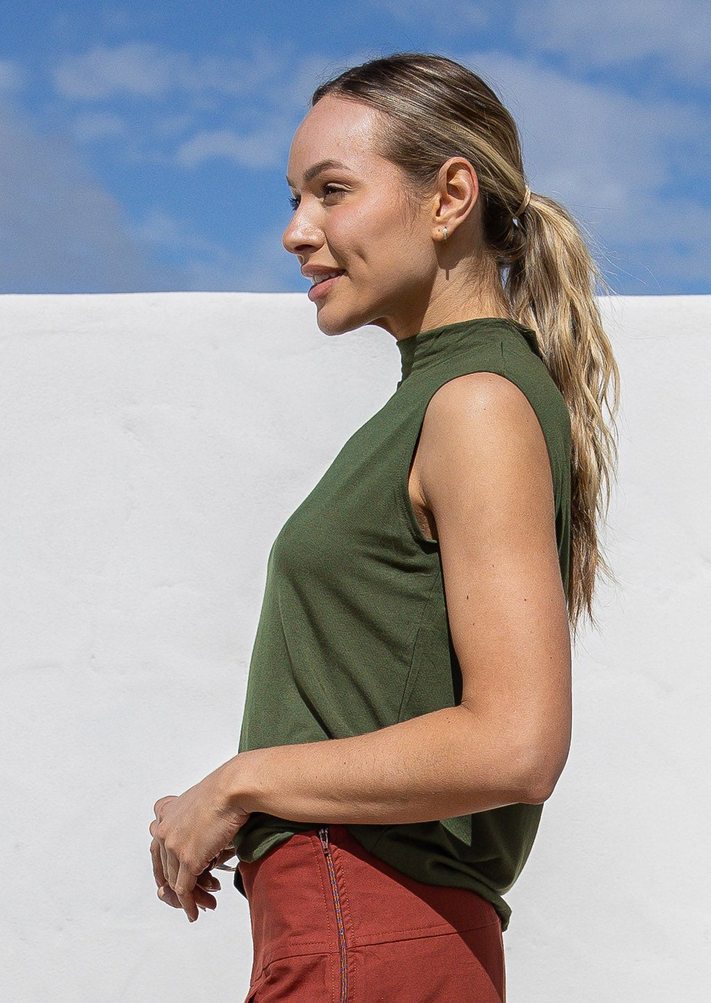 Woman wearing an olive green sleeveless top against a white wall with blue sky.