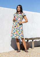 Woman wearing a pastel print cotton knee length day dress standing against a white wall with a blue sky.