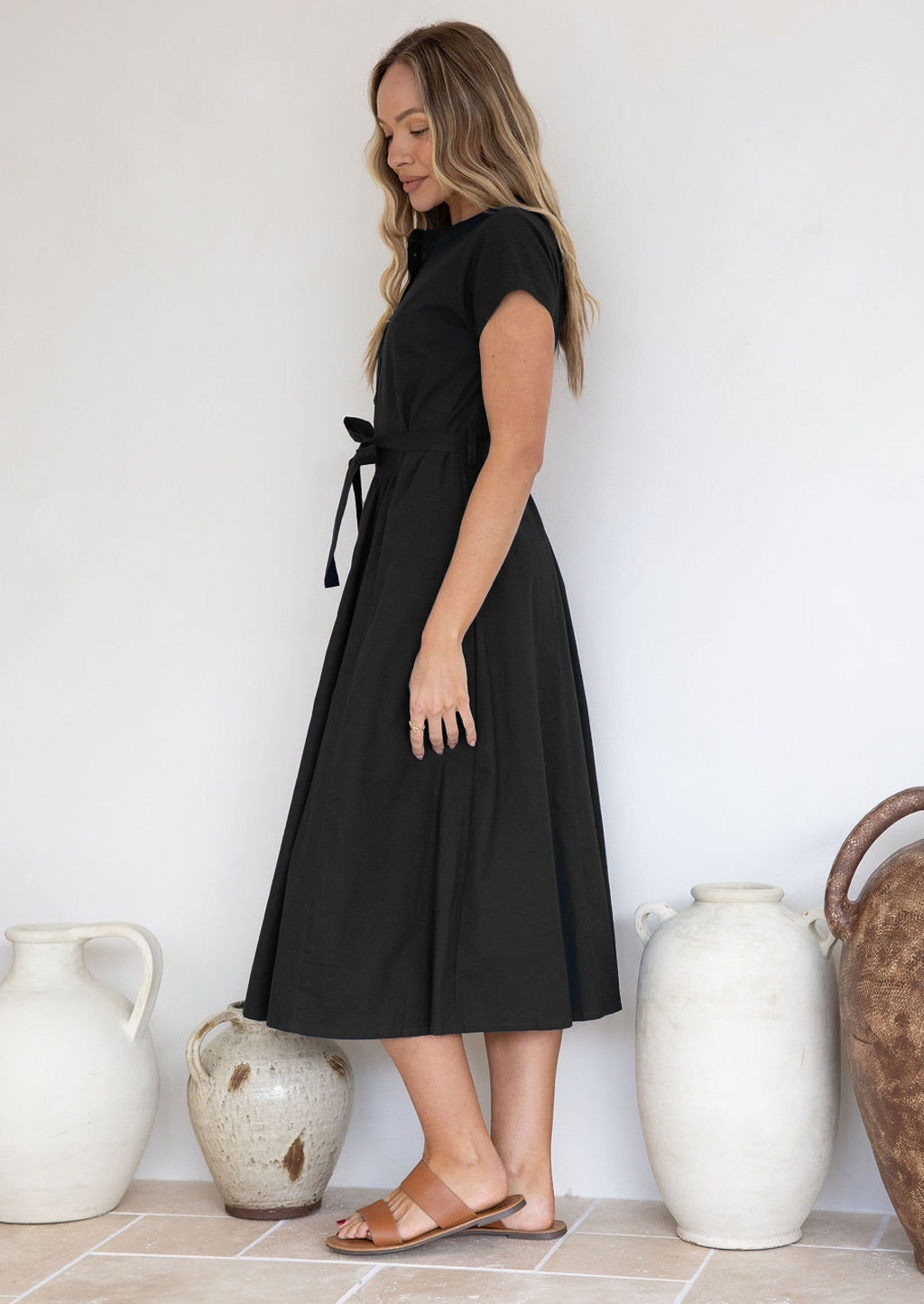 Woman standing side on, wearing a black 50's sty;e cotton dress with cap sleeves and a waist tie