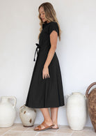 Woman standing side on, wearing a black 50's sty;e cotton dress with cap sleeves and a waist tie