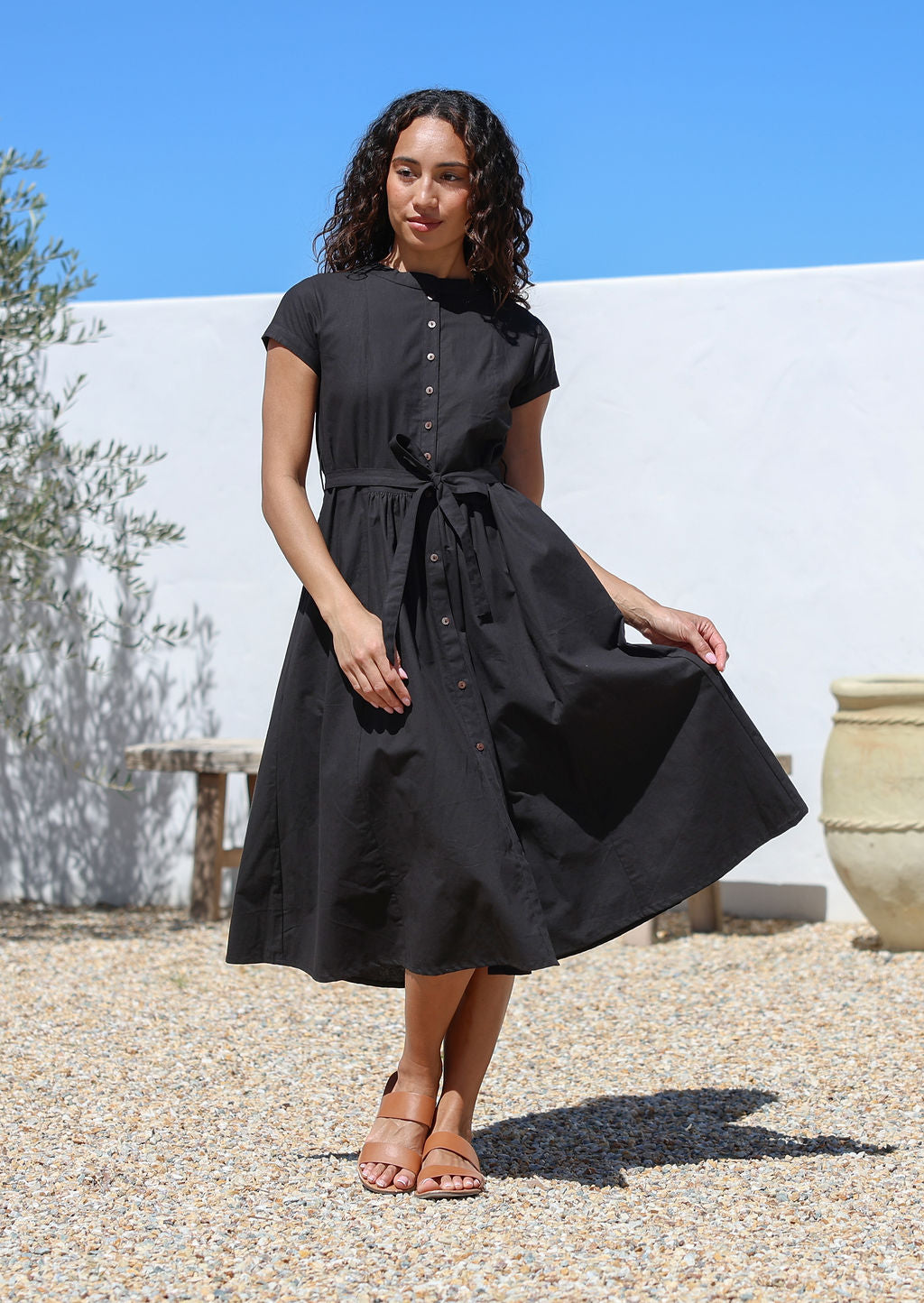 Woman wearing a black cotton retro style dress standing outdoors with a white wall 