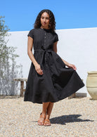 Woman wearing a black cotton retro style dress standing outdoors with a white wall 