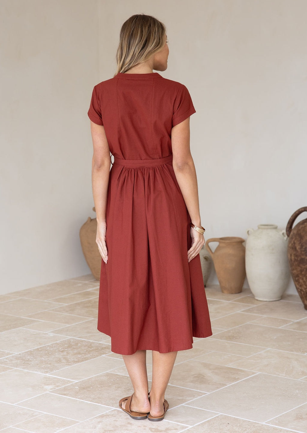 Model wears a 100% cotton rust-colored midi-length dress with a high neckline, pockets, and a removable waist tie 
