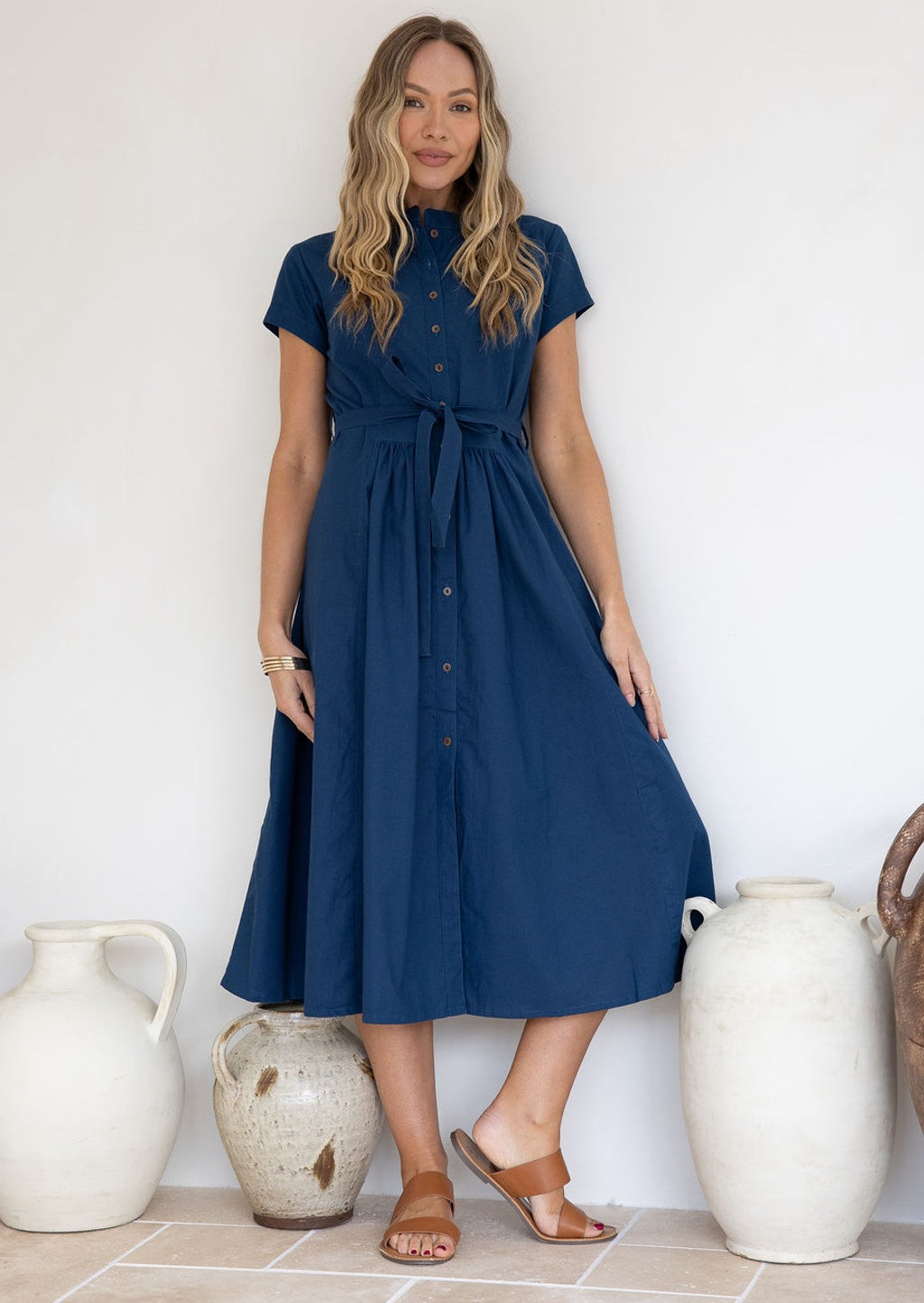 Woman wearing a 50's style cotton blue dress standing against a white wall with decorative vases