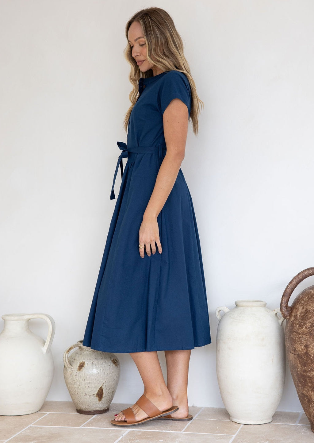 Model stands on side wearing a deep blue 50's style midi length cotton dress with cap sleeves and waist tie