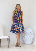 Woman wearing a pink and green floral print on a blue base sleeveless dress with hands in side pockets