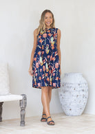 Woman standing in a pink and green native floral print on a blue base sleeveless dress 