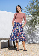 Woman wearing a dusty rose top with floral below knee length, navy cotton skirt with hands in pocket.