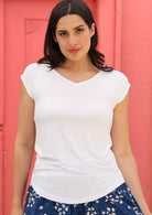 Woman with dark hair wearing a white v-neck short cap sleeve rayon top in front of a pink wall.