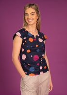 Woman wears a cap sleeve cotton top with a keyhole cutout in the neckline, in a blue based multi coloured dotty print