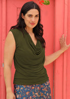 Woman standing in front of a pink door wearing a cowl neck rayon olive cap sleeve top.