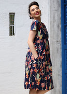 Model wears colorful native pink and green floral printed 100% cotton knee length dress with hands in pocket.