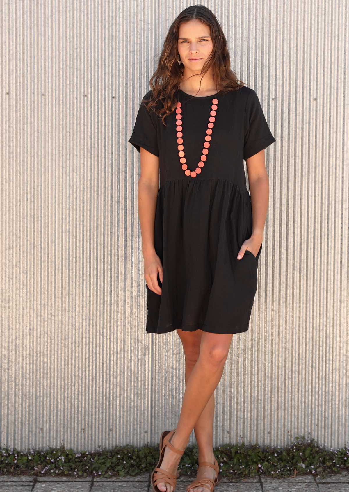 Woman in knee length black dress with orange necklace and one hand in pocket 
