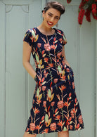 Woman wears Cap Sleeved 100% Cotton Dress in pink and green floral print on a blue base, with hands in Pockets